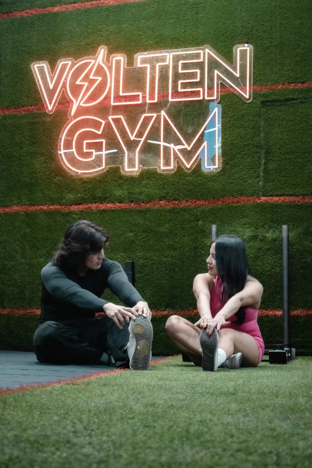 Dos socios estirando sobre pasto sintético bajo el neón de Volten Gym.
